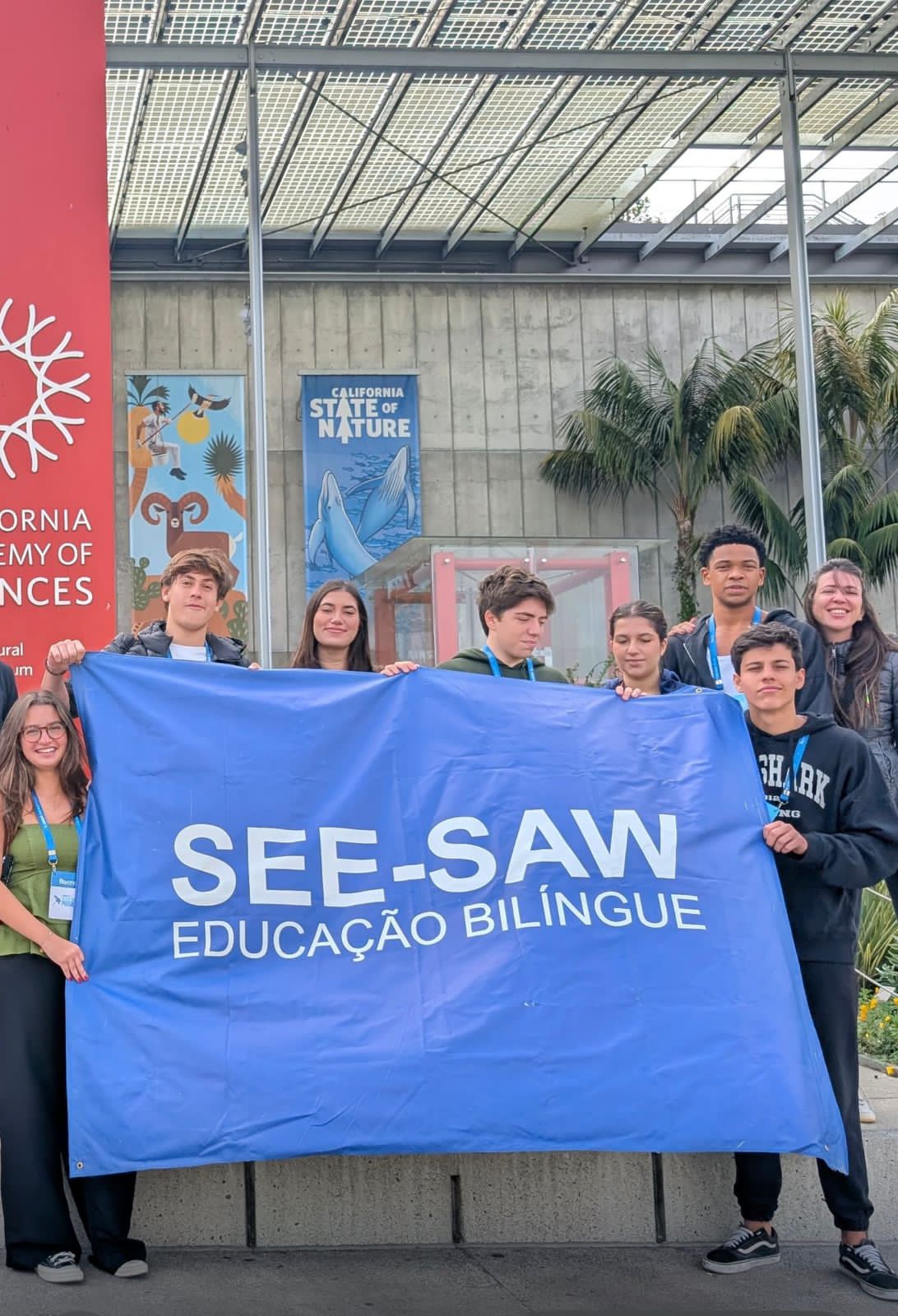 Grupo de alunos sorridentes da SEE-SAW Educação Bilíngue segurando uma faixa azul com o logo da escola em frente a um prédio moderno na Califórnia durante experiência acadêmica no Vale do Silício